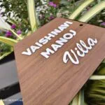 Wooden name board with raised white 3D letters reading Vaishnavi Manoj Villa