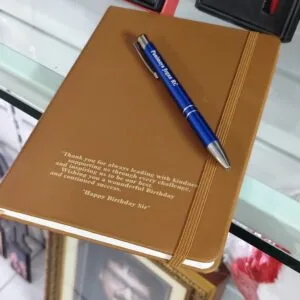 Tan hardbound notebook engraved with a message beside a personalised blue pen